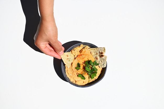 Foto vista superior de una mano sumergiendo pan de pita en un plato vegano de consistencia cremosa hummus oriental con perejil en un tazón