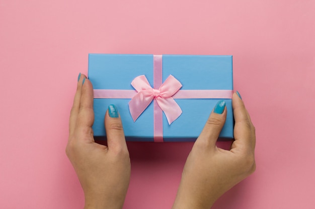 Vista superior manos femeninas con caja de regalo azul con lazo rosa festivo sobre un fondo rosa suave