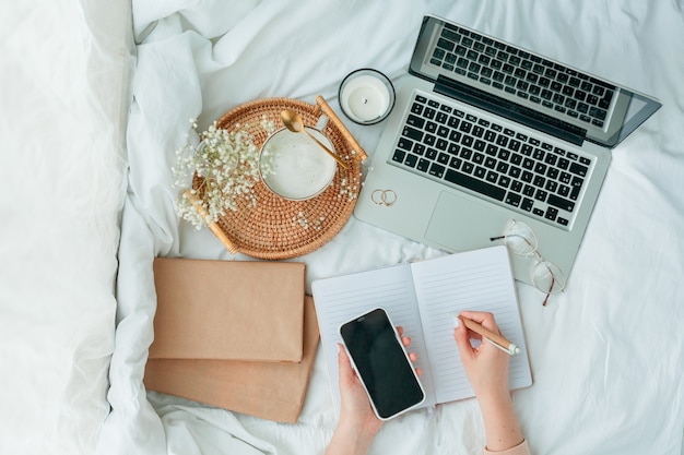 Vista superior de manos femeninas escribiendo en un cuaderno con un capuchino en bandeja de mimbre, un teléfono móvil y una computadora portátil alrededor