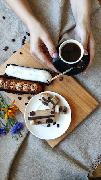 Vista superior en manos femeninas con flores de verano cerca de una taza de café negro y servido eclairs