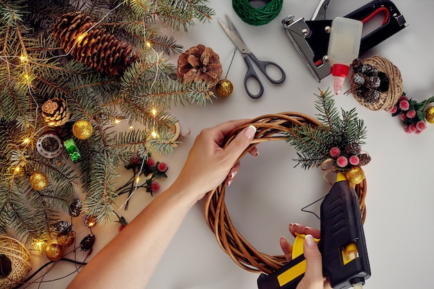 Vista superior de manos femeninas hacen una corona de Navidad. Regalos empaquetados y pergaminos, ramas de abeto y herramientas en el cuadro blanco. Preparándose para las vacaciones de Navidad o Año Nuevo
