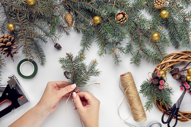 Vista superior de manos femeninas hacen una corona de Navidad. Regalos empaquetados y pergaminos, ramas de abeto y herramientas en el cuadro blanco. Preparándose para las vacaciones de Navidad o Año Nuevo