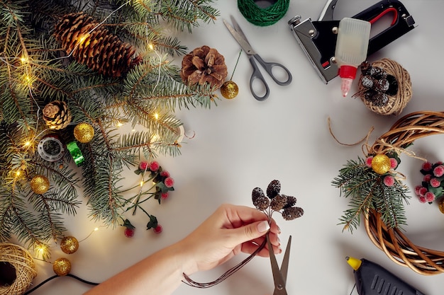 Vista superior de manos femeninas hacen una corona de Navidad. Regalos empaquetados y pergaminos, ramas de abeto y herramientas en el cuadro blanco.