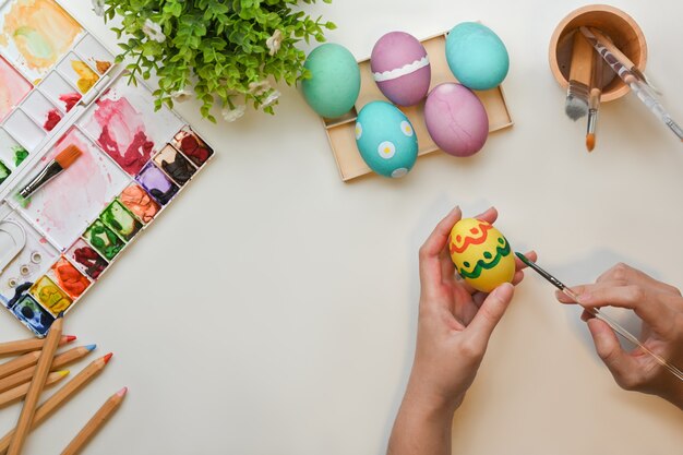 Vista superior de manos femeninas pintando huevos para prepararse para el festival de Pascua en mesa de manualidades con herramientas de pintura