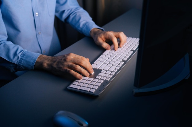 Vista superior de las manos del hombre irreconocible que usa la computadora escribiendo en el teclado inalámbrico en la habitación oscura