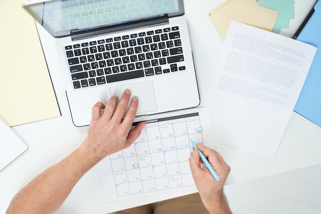 Vista superior de las manos del hombre usando su computadora portátil y marcando los días en el calendario mientras