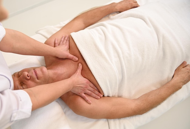 Vista superior de las manos de una masajista profesional que hace masaje manual de rehabilitación relajante de cuello y hombros a un hombre caucásico maduro relajante y guapo, paciente en clínica de bienestar