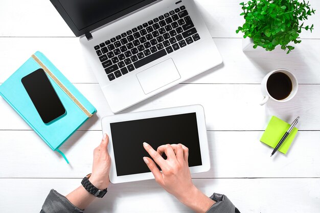 Vista superior de las manos de la mujer escribiendo en el teclado de la computadora portátil colocada en el escritorio de la oficina blanca con teléfono inteligente en blanco, taza de café, plantas decorativas y suministros. Bosquejo