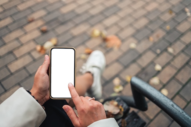 Vista superior de las manos de una mujer sosteniendo un teléfono inteligente con pantalla en blanco en blanco. Copie el espacio en el jardín de otoño. Mujer leyendo texto y viendo videos en un teléfono móvil