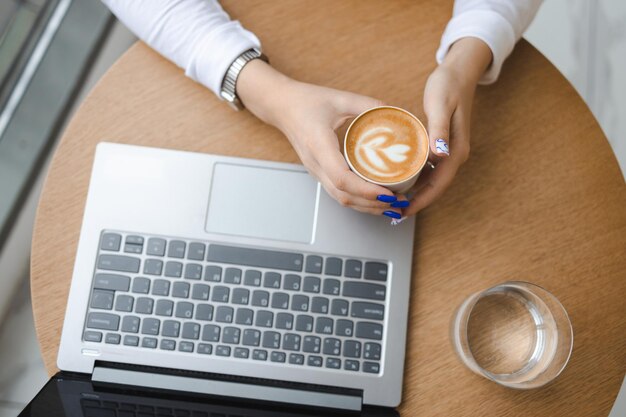 Vista superior de las manos de la mujer con una taza de café sentada en un café con una computadora portátil