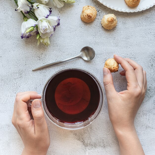Vista superior de las manos de una mujer con una taza de té, galletas y flores.
