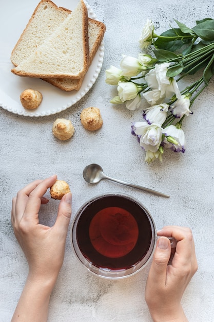 Vista superior de las manos de una mujer con una taza de té, galletas, pan tostado y flores.