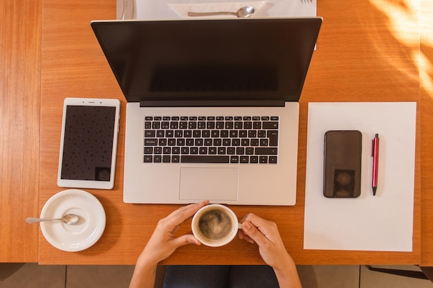 Vista superior de las manos de una niña sosteniendo un café con sus objetos tecnológicos a su alrededor