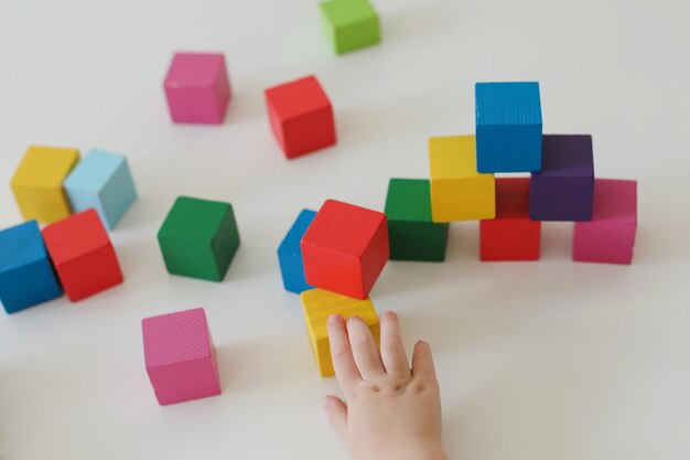 Vista superior de las manos del niño jugando y construyendo con coloridos ladrillos de juguete de madera en la mesa de madera blanca