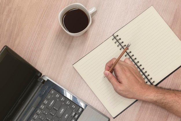 Vista superior mão segurando o lápis e escrevendo no caderno com laptop e xícara de café preta na mesa