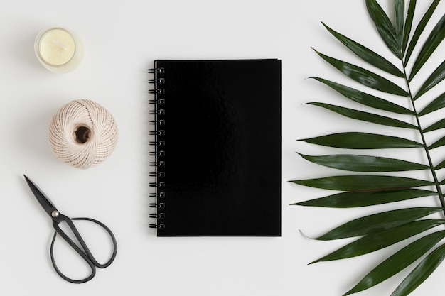 Vista superior de una maqueta de cuaderno negro con accesorios de espacio de trabajo y una hoja de palma sobre una mesa blanca