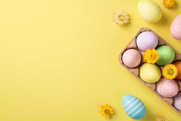 Vista superior de medio cartón de papel con huevos y flores multicolores de Pascua y tres huevos pastel y flores brillantes en el fondo vacío amarillo aislado