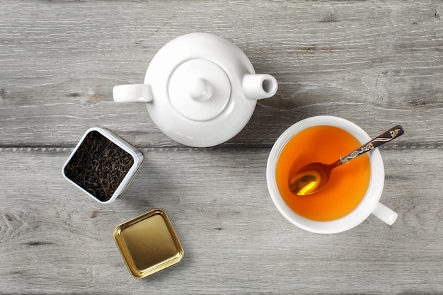 Vista superior de la mesa: carrito de té de latón, tetera de porcelana blanca y taza de cerámica con cuchara de plata y bebida de ámbar caliente, sobre un escritorio de madera gris.