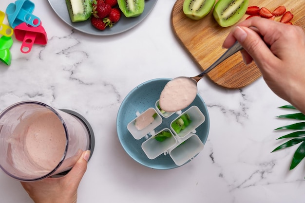 Vista superior de la mesa de la cocina con manos femeninas vertiendo helado en moldes Hacer helado casero de yogur, fresas y kiwi