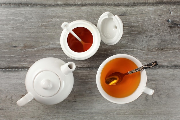 Vista superior de la mesa: escritorio de madera gris con tetera, taza de té con bebida caliente, cuchara de plata y tarro pequeño de porcelana blanca.