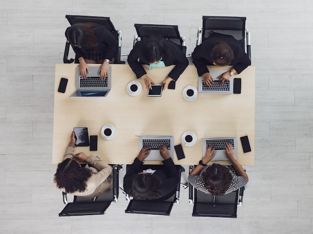 Vista superior de la mesa de madera de la conferencia de reuniones con seis mujeres empresarias ejecutivas sentadas en cada una de las sillas con teléfonos móviles, portátiles y tabletas en la sala de reuniones.