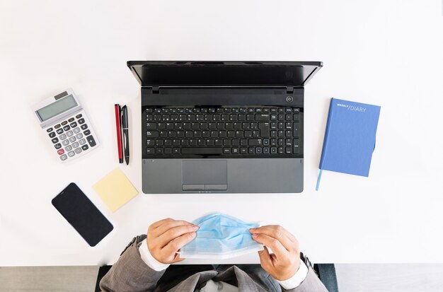 Vista superior de una mesa de oficina blanca con una computadora portátil, calculadora, teléfono celular, poste, bolígrafos, cuaderno y con un trabajador de negocios con una mascarilla debido a la pandemia de coronavirus covid19