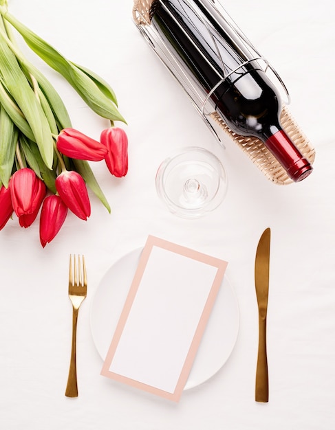 Vista superior de la mesa con tarjeta de menú, cubiertos, tulipanes rojos frescos y vino sobre un mantel de tela blanca