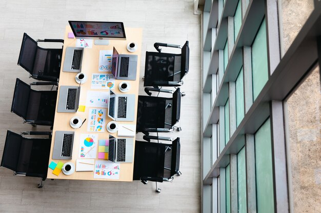 Foto vista superior de la mesa de trabajo de la sala de reuniones en la oficina de la empresa vacía llena de computadoras portátiles, tazas de café, informes, documentos de papeleo, sillas negras y tablero de vidrio de pantalla de monitor en blanco cerca de las ventanas.