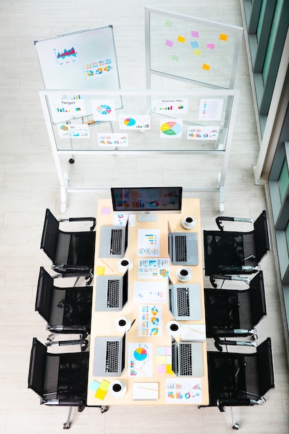 Vista superior de la mesa de trabajo de la sala de reuniones en la oficina de la empresa vacía llena de computadoras portátiles, tazas de café, informes, documentos de papeleo, sillas negras y tablero de vidrio de pantalla de monitor en blanco cerca de las ventanas.