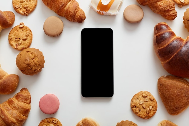 Foto vista superior de la mesa con varias galletas, pasteles, cruasanes, panecillos, don teléfono inteligente de fondo blanco con espacio de copia de pantalla vacía para el área de publicidad de promociones