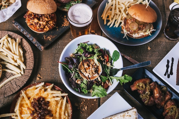 Vista superior de la mesa con variedad de platos, hamburguesas, papas fritas y ensalada, bebidas, alitas de pollo y salsa en la mesa de madera. Menú del restaurante.