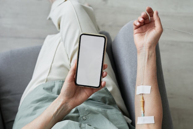 Vista superior mínima cerca de una mujer irreconocible recibiendo goteo intravenoso y usando un teléfono inteligente con pantalla en blanco, espacio de copia