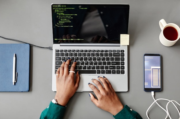 Vista superior mínima de uma mulher negra irreconhecível usando laptop e escrevendo código na mesa cinza do local de trabalho