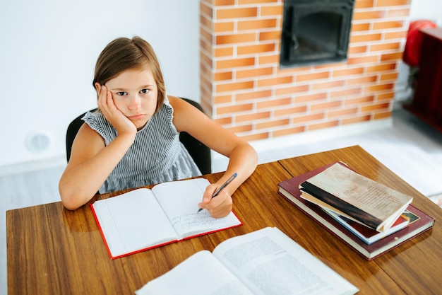 Foto vista superior mirando a la cámara insatisfecho hermoso escolar se sienta en la mesa sosteniendo la cabeza con la mano extraña los libros de texto en frente el niño está cansado de las tareas las lecciones aburridas sin interés espacio de copia