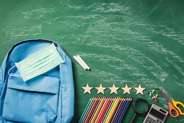 Foto vista superior de una mochila escolar azul en una pizarra verde con protección de máscara facial