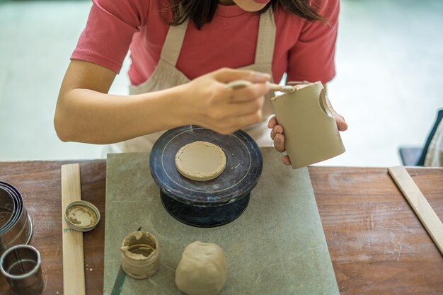 Vista superior Mujer alfarera trabajando en la rueda de alfarero haciendo vasijas de cerámica de arcilla en el taller de cerámica Enfoque mano mujer joven adjuntando parte del producto de arcilla al futuro producto de cerámica Taller de cerámica