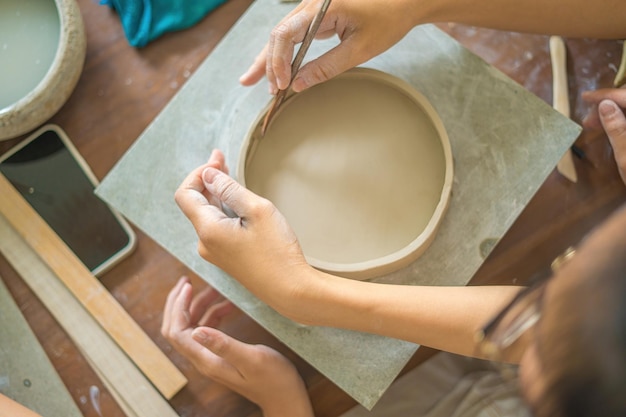 Vista superior Mujer alfarera trabajando en la rueda de alfarero haciendo vasijas de cerámica de arcilla en el taller de cerámica Enfoque mano mujer joven adjuntando parte del producto de arcilla al futuro producto de cerámica Taller de cerámica