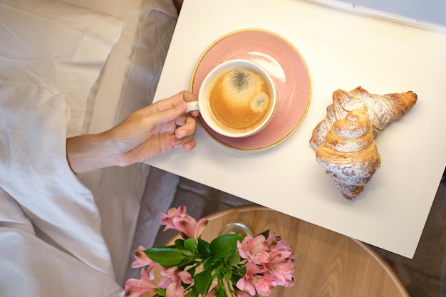 Foto vista superior de la mujer de la cosecha acostada en una cama blanca suave cubierta de edredón y extendiendo la taza de café y los croissants colocados en la mesa blanca en el dormitorio en casa