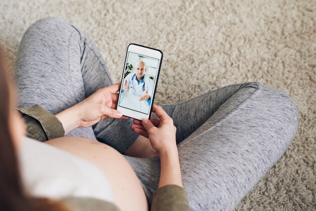 Vista superior de una mujer embarazada sentada con las piernas cruzadas en el piso y usando un teléfono inteligente mientras habla con el ginecólogo a través del chat de video