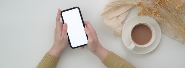 Vista superior de una mujer freelance usando un teléfono inteligente de pantalla en blanco en la mesa del desayuno