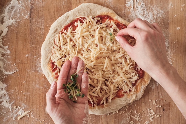 Vista superior de una mujer irreconocible que agrega hierbas a la pizza cruda colocada en una superficie de madera con harina