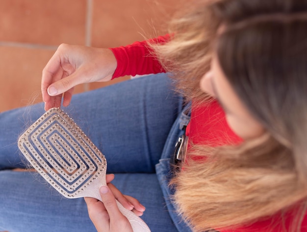 Vista superior de una mujer irreconocible sentada y sosteniendo un cepillo de pelo lleno de pelo en otoño con traje rojo y de mezclilla