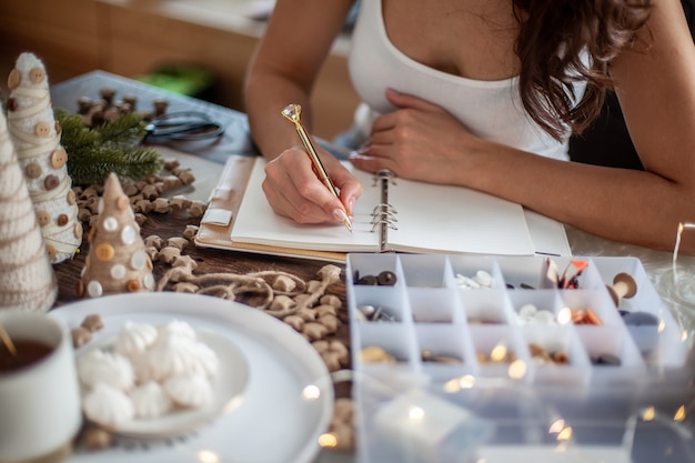 La vista superior de la mujer joven está escribiendo metas para el año nuevo 2021 en un diario en el escritorio con adornos de árboles de Navidad hechos a mano, estrellas, botones y té con malvavisco. Concepto de planificación de nuevos objetivos.