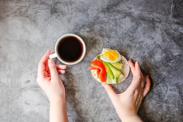 Vista superior de la mujer joven que tiene una pausa para el almuerzo. Tortitas de arroz crujiente con aguacate y salmón fresco salado. Manos masculinas con alimentos ricos en grasas y proteínas y una taza de café. Espacio para texto.