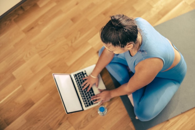 Vista superior de una mujer joven que usa una computadora portátil mientras hace entrenamiento físico en casa.