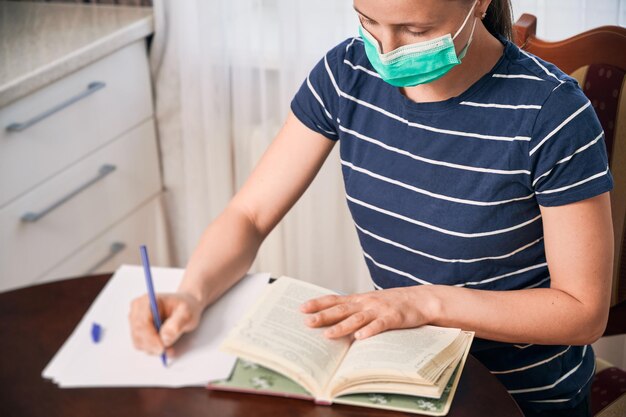 Foto vista superior de una mujer joven sentada en la mesa con una máscara leyendo un libro tomando notas estudio universitario remoto en casa distancia social crisis de cuarentena global covid19 enfoque selectivo