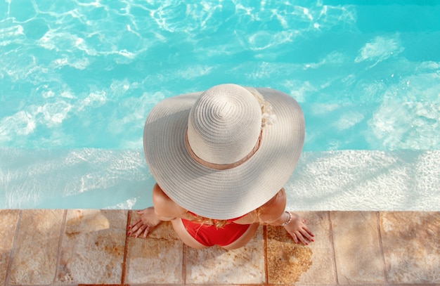 Vista superior de la mujer joven en traje de baño rojo de una pieza y sombrero de paja relajarse cerca de una piscina con las piernas en el agua