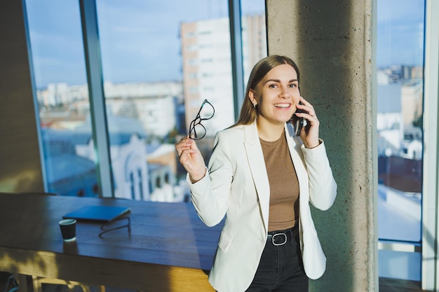 Vista superior de una mujer positiva con ropa informal parada cerca de una ventana grande y hablando por teléfono dando información durante el proyecto remoto Espacio de trabajo moderno para trabajo remoto