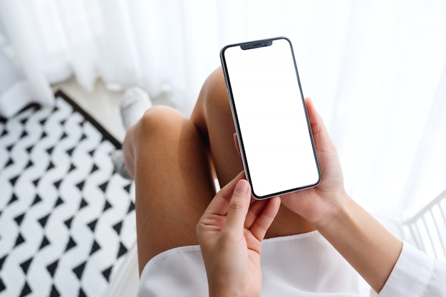 Vista superior de la mujer que sostiene el teléfono móvil con pantalla blanca de escritorio en blanco mientras está sentado en el dormitorio con sensación de relajación