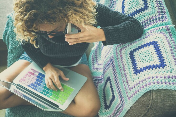 Foto vista superior de la mujer que trabaja en la computadora portátil con un teclado colorido mientras asiste a una llamada de teléfono móvil relajándose en el sofá en casa. mujer que usa la computadora portátil y el teléfono móvil mientras está sentado con las piernas cruzadas en un cómodo sofá